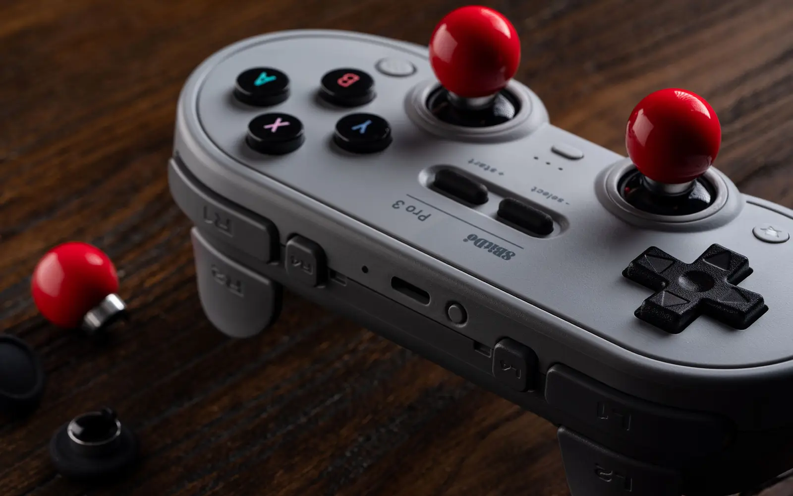 Gray 8BitDo Pro 3 controller on a wooden desk with red ball top joystick caps