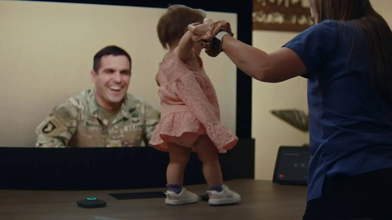 Toddler taking steps in front of a screen showing a smiling soldier