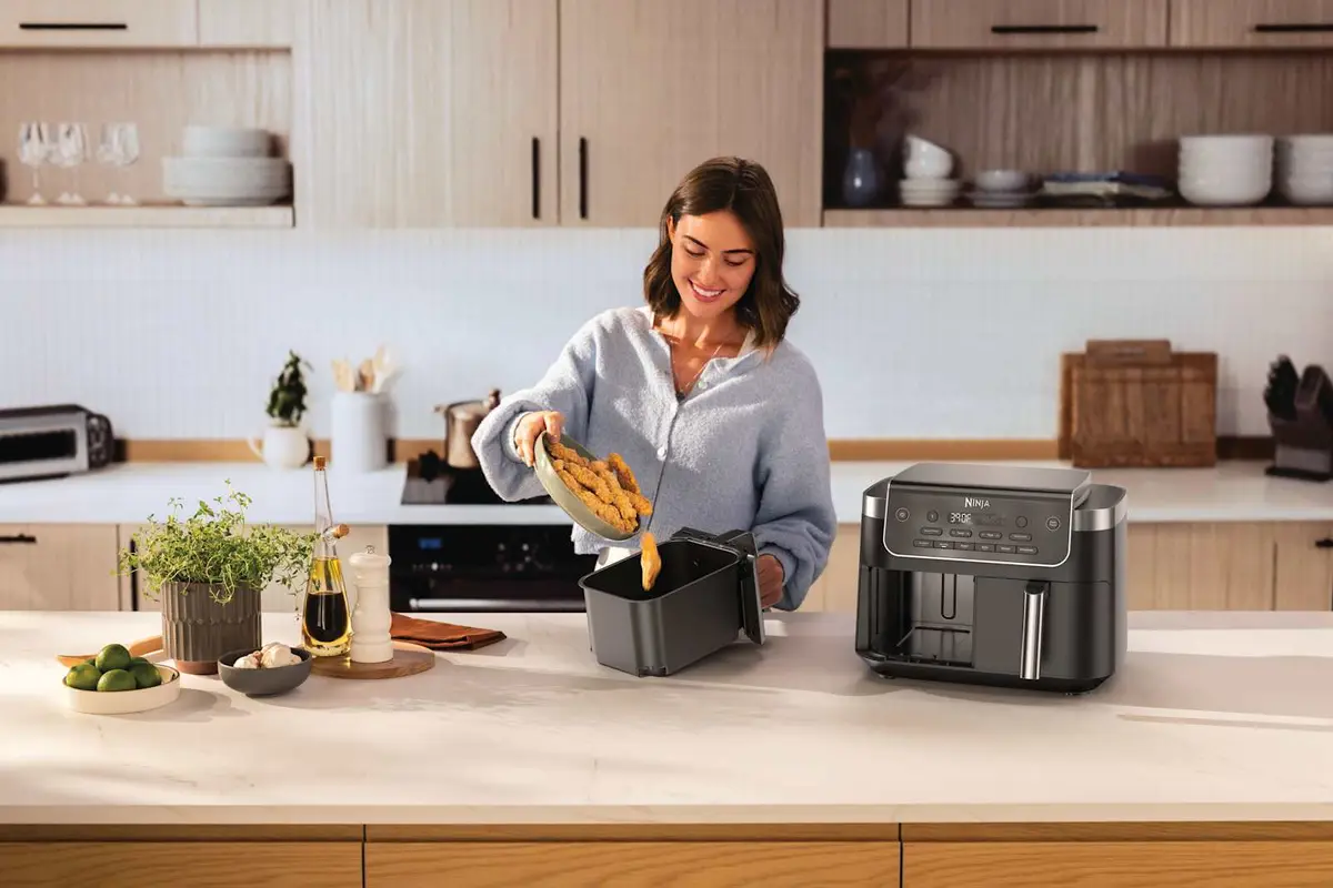 Person tipping cooked food from a basket next to a Ninja DualZone air fryer