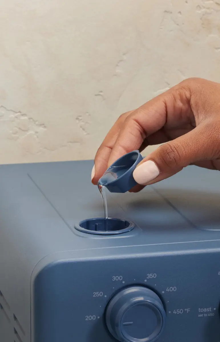 A hand pours water into the top steam opening of a blue Wonder Oven