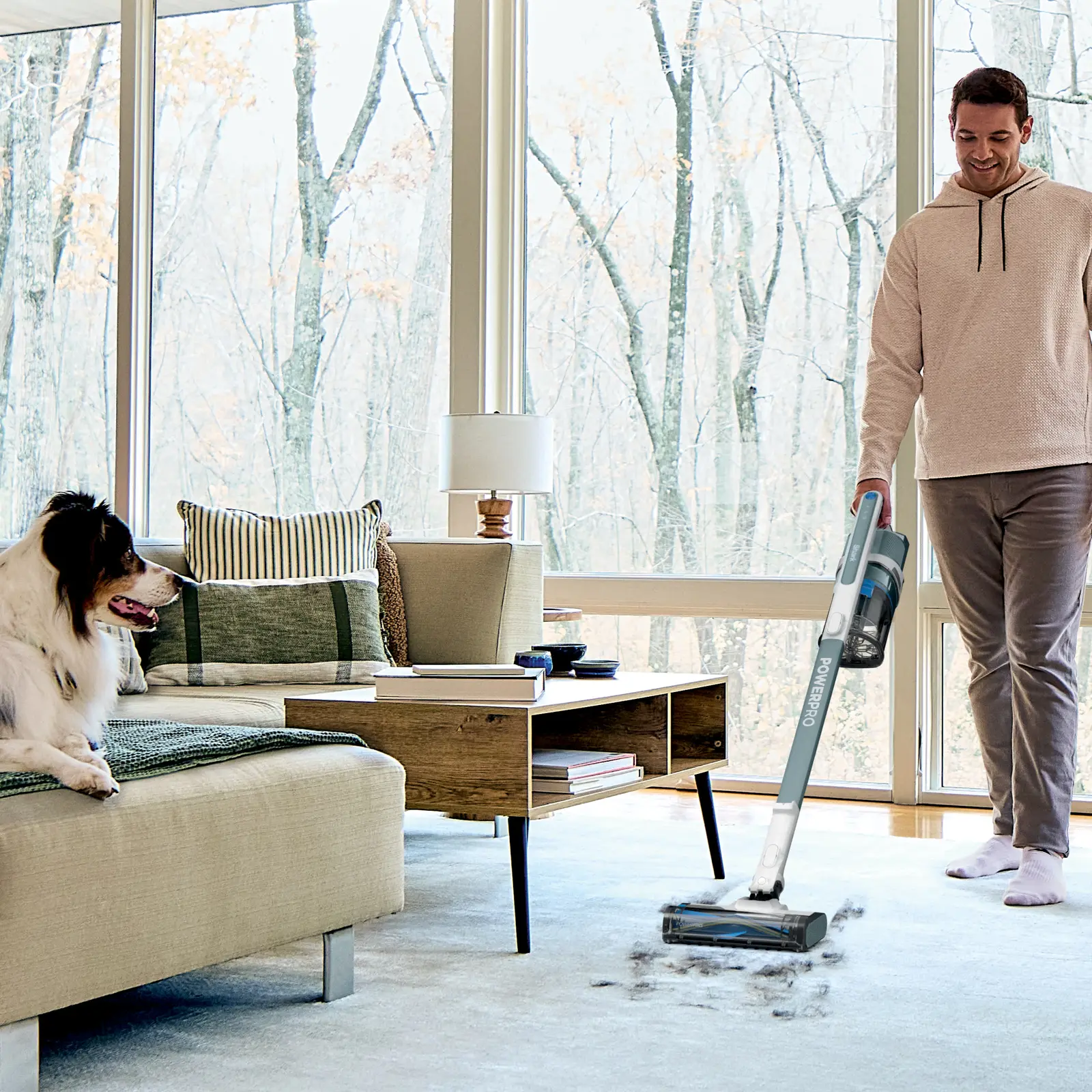 Man using a Shark PowerPro Plus cordless stick vacuum on a rug in a living room with a dog